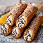 Plated cannoli recipe pastries filled with creamy ricotta filling, dusted with powdered sugar and drizzled with chocolate, served with a lemon wedge and mint garnish.