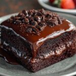 A close-up of a single slice of rich, moist chocolate cake recipe topped with glossy ganache and chocolate shavings on a grey plate.