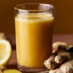A clear glass filled with a bright golden-yellow ginger shot recipe, surrounded by pieces of fresh ginger root and a halved lemon on a warm-toned surface.