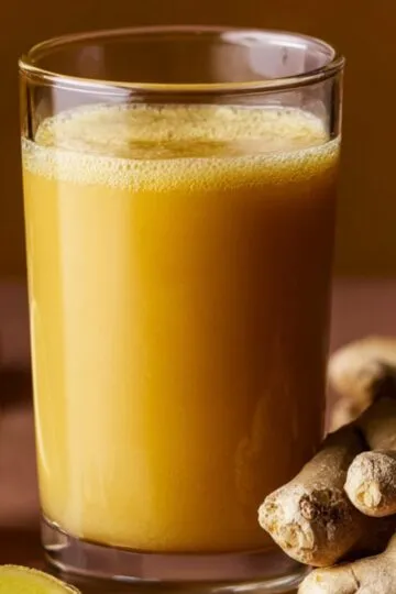 A clear glass filled with a bright golden-yellow ginger shot recipe, surrounded by pieces of fresh ginger root and a halved lemon on a warm-toned surface.