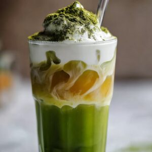 A tall glass of iced matcha latte showcasing a vivid green matcha layer at the bottom, a golden honey swirl in the middle, and frothy white milk foam on top dusted with matcha powder, served with a clear straw.