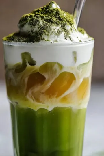 A tall glass of iced matcha latte showcasing a vivid green matcha layer at the bottom, a golden honey swirl in the middle, and frothy white milk foam on top dusted with matcha powder, served with a clear straw.