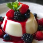 A creamy white panna cotta recipe unmolded on a white plate, drizzled with vivid red berry sauce that pools at its base. The top is crowned with a cluster of raspberries, blackberries, and blueberries, and finished with a sprig of vibrant green mint.