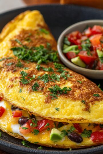 Folded golden omelette recipe on a black plate, filled with melted cheese, black beans, diced tomatoes, and avocado, garnished with chopped parsley and served alongside a small bowl of fresh pico de gallo.