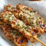 Close‑up of three Ranch Garlic Parmesan Chicken Skewers on parchment paper, each piece of chicken coated in a golden‑brown crust of Parmesan cheese and herbs, with flecks of chopped parsley and a light char for a smoky finish