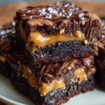 Two rich, fudgy Snickers Brownies squares layered with gooey caramel and chopped peanuts, drizzled with melted chocolate and sprinkled with sea salt, presented on a light-colored plate.