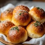 Basket of golden, shiny Bread Rolls Recipe sprinkled with coarse salt and herbs, resting on a white cloth.