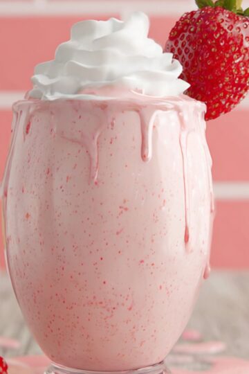 A creamy pink strawberry milkshake in a footed glass, topped with swirled whipped cream and garnished with a whole strawberry on the rim. The shake has begun to drip down the sides onto the wooden table, surrounded by scattered fresh strawberries and a blurred bottle in the background against a pink-tiled wall.”