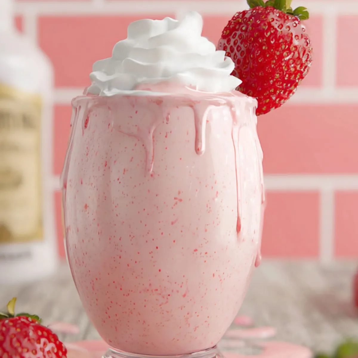A creamy pink strawberry milkshake in a footed glass, topped with swirled whipped cream and garnished with a whole strawberry on the rim. The shake has begun to drip down the sides onto the wooden table, surrounded by scattered fresh strawberries and a blurred bottle in the background against a pink-tiled wall.”