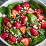 A white bowl filled with fresh baby spinach leaves, halved ripe Strawberry Spinach Salad, crumbled feta cheese, candied pecans, and a few blueberries, all lightly coated in vinaigrette.