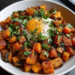 A white bowl filled with golden‑orange sweet potato hash cubes, their edges crisped and caramelized, scattered with fresh chopped parsley, and topped with a perfectly runny sunny‑side‑up egg, all bathed in soft natural light.