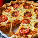 A close‑up view of a baked tomato pie in a white ceramic dish with one wedge removed: the flaky golden crust holds layers of juicy tomato slices and melted cheese that stretches as the slice is lifted, and the top is sprinkled with fresh herbs.
