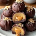 A stack of rich, homemade buckeyes recipe truffles with glossy dark chocolate coating, topped with crushed cookie crumbs. The center of one cut-open truffle reveals a smooth peanut butter filling surrounded by chocolate. The caramel-like center is soft and gooey, highlighting the decadent texture.