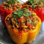 Three colorful stuffed bell peppers (yellow and red) on a plate, each filled with a savory rice and tomato mixture and garnished with chopped fresh herbs.