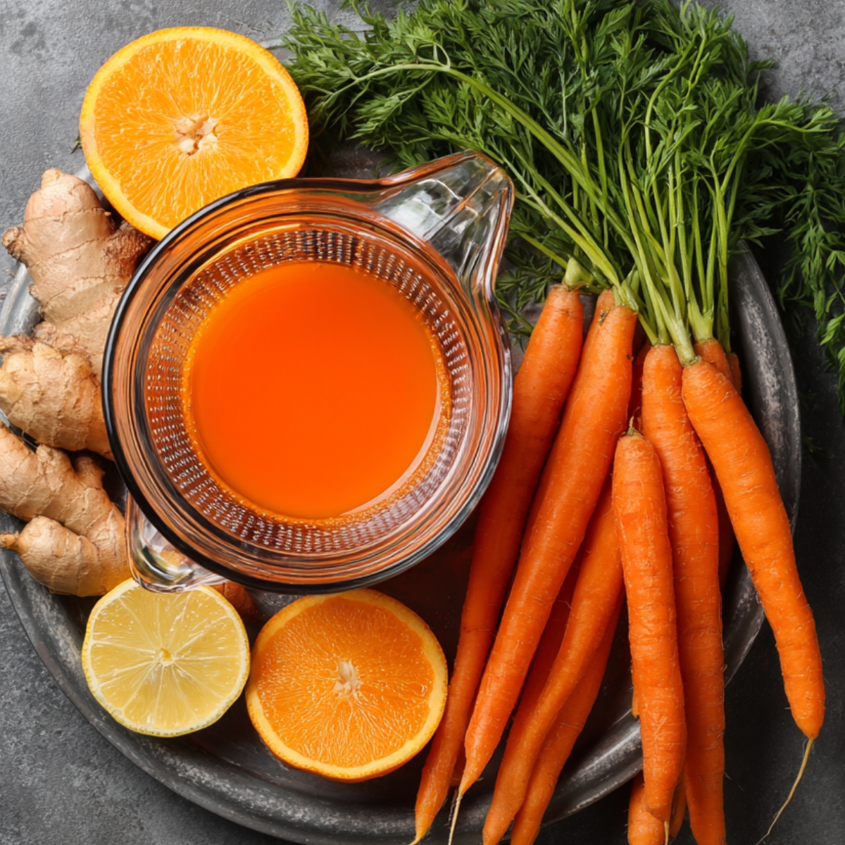 A top-down view of a dark stone platter filled with fresh juicing ingredients: a glass pitcher of bright orange carrot juice at the center, a bundle of vibrant young carrots with green tops to the right, pieces of fresh ginger to the left, and halved citrus fruits (lemon and orange) at the top and bottom. The rustic stone surface beneath adds a farmhouse kitchen feel, and soft natural light highlights the vivid colors and textures of each element.