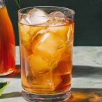 A tall, clear glass filled with amber-colored iced tea and large ice cubes, garnished with a fresh lime wedge on the rim and a cinnamon stick straw; in soft focus behind it sits a glass pitcher of more iced tea, all set against a deep blue background.