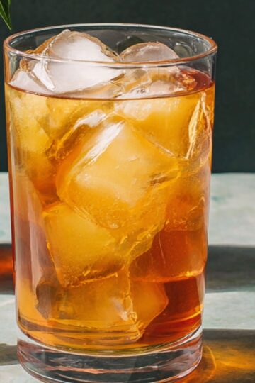 A tall, clear glass filled with amber-colored iced tea and large ice cubes, garnished with a fresh lime wedge on the rim and a cinnamon stick straw; in soft focus behind it sits a glass pitcher of more iced tea, all set against a deep blue background.