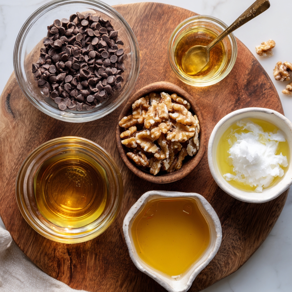 A wooden round cutting board holding clear and ceramic bowls filled with ingredients for sweet potato brownies. Visible are dark chocolate chips in a glass bowl, chopped walnuts in a small wooden bowl, white coconut oil in a cream ceramic dish, and golden maple syrup in both a small jar and a unique pouring bowl. Soft natural daylight and a marble counter in the background add to the homemade kitchen feel.