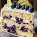 A thick slice of two-layer lemon blueberry cake on a cream-colored ceramic plate, with a silver dessert fork resting beside it. The golden yellow sponge is studded with juicy blueberries and filled with a creamy white frosting. More frosting coats the top, where fresh blueberries and thin lemon slices are artfully arranged and lightly dusted with powdered sugar. In the blurred background, warm wooden tones suggest a cozy home kitchen setting.