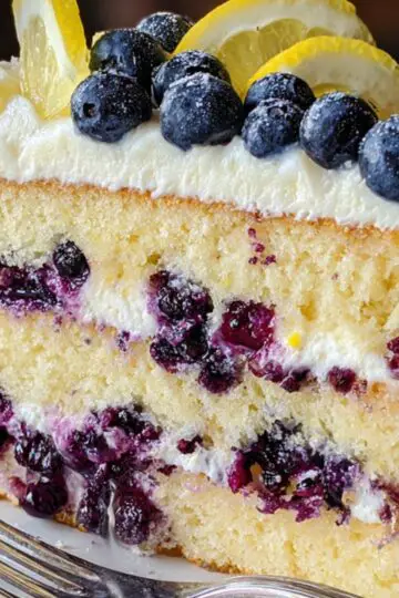 A thick slice of two-layer lemon blueberry cake on a cream-colored ceramic plate, with a silver dessert fork resting beside it. The golden yellow sponge is studded with juicy blueberries and filled with a creamy white frosting. More frosting coats the top, where fresh blueberries and thin lemon slices are artfully arranged and lightly dusted with powdered sugar. In the blurred background, warm wooden tones suggest a cozy home kitchen setting.