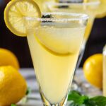 A close-up of a homemade lemon drop martini in a tall, conical glass with a crystalline sugar rim. The pale yellow cocktail is chilled tiny beads of condensation cling to the glass and a thin wheel of lemon floats inside, while another lemon slice perches on the rim. In soft focus behind it, whole lemons and fresh green herb leaves rest on a light marble tabletop, giving the scene a bright, fresh, and inviting feel.