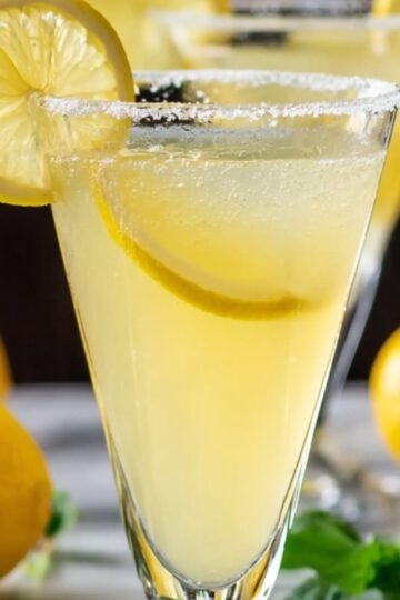 A close-up of a homemade lemon drop martini in a tall, conical glass with a crystalline sugar rim. The pale yellow cocktail is chilled tiny beads of condensation cling to the glass and a thin wheel of lemon floats inside, while another lemon slice perches on the rim. In soft focus behind it, whole lemons and fresh green herb leaves rest on a light marble tabletop, giving the scene a bright, fresh, and inviting feel.