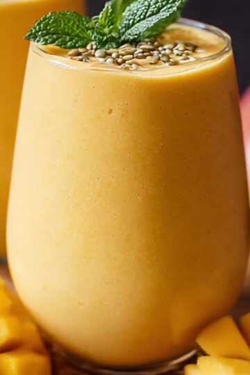 Homemade mango smoothie in a clear glass, topped with mint and seeds, surrounded by fresh mango cubes on a wooden surface.