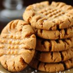 A stack of thick, golden-brown homemade peanut butter cookies with crisscross fork marks and a light sugar sprinkle, sitting on a plate in warm, cozy lighting.