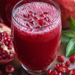 A tall glass of freshly pressed pomegranate juice topped with a handful of ruby-red pomegranate arils, set on a rustic wooden tabletop beside a halved pomegranate, scattered seeds, and a few glossy green leaves; soft, natural light highlights the deep crimson color and the tiny bubbles on the juice surface