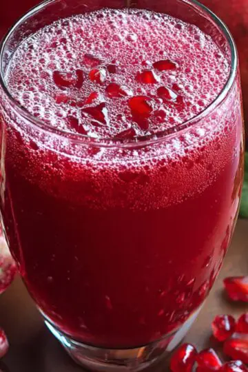 A tall glass of freshly pressed pomegranate juice topped with a handful of ruby-red pomegranate arils, set on a rustic wooden tabletop beside a halved pomegranate, scattered seeds, and a few glossy green leaves; soft, natural light highlights the deep crimson color and the tiny bubbles on the juice surface