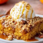 A warm, homemade slice of Pumpkin Dump Cake Recipe served on a ceramic plate, topped with a generous scoop of vanilla ice cream. The cake has a golden-orange pumpkin base, crumbly brown sugar streusel topping, and visible toasted pecans. Thick caramel sauce is drizzled over the dessert and pooling on the plate. In the softly blurred background, small decorative pumpkins, a glass of milk, and rust-colored napkins evoke a cozy fall setting.