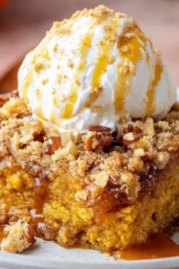 A warm, homemade slice of Pumpkin Dump Cake Recipe served on a ceramic plate, topped with a generous scoop of vanilla ice cream. The cake has a golden-orange pumpkin base, crumbly brown sugar streusel topping, and visible toasted pecans. Thick caramel sauce is drizzled over the dessert and pooling on the plate. In the softly blurred background, small decorative pumpkins, a glass of milk, and rust-colored napkins evoke a cozy fall setting.