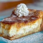 A homemade slice of sopapilla cheesecake with a golden caramelized top, creamy filling, flaky crust, dusted with powdered sugar, and topped with whipped cream on a blue plate.
