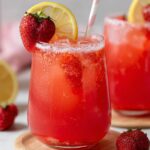 Two glasses of homemade strawberry lemonade with ice, fresh strawberries, lemon slices, and striped straws, set on wooden coasters. A halved lemon, a strawberry, a pink napkin, and a blurred pitcher with flowers appear in the background.