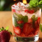 A short clear glass filled with roughly crushed ice, fresh sliced strawberries pressed against the walls, and whole mint leaves muddled throughout, topped with a mint sprig. Tiny bubbles from sparkling water cling to the fruit and herbs, and a whole strawberry mojito rests beside the glass on a warm wooden surface, with a softly blurred background evoking a homemade summer cocktail vibe.
