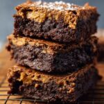 A clear glass mixing bowl on a marble countertop filled with smooth, bright orange mashed Sweet Potato Brownies. A metal whisk rests inside the bowl, and the mixture looks creamy and freshly stirred, lit by soft daylight.