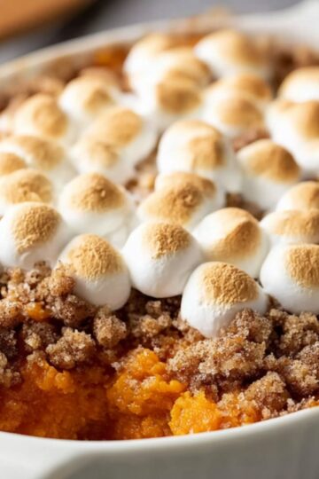 A close-up of a homemade sweet potato casserole in a white ceramic baking dish. The vibrant orange sweet potato mash peeks through where a scoop was taken, beneath a golden-brown pecan-oat streusel layer and perfectly toasted mini marshmallows with light tan tops. The edges of the dish show bits of melted marshmallow and crumb topping, all lit by warm, natural kitchen light.
