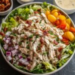 Bowl of creamy Chicken Salad Recipe on a bed of romaine lettuce, topped with shredded chicken, diced red onion, halved cherry tomatoes, and bacon bits, with a small ramekin of dressing and extra bacon in the background.