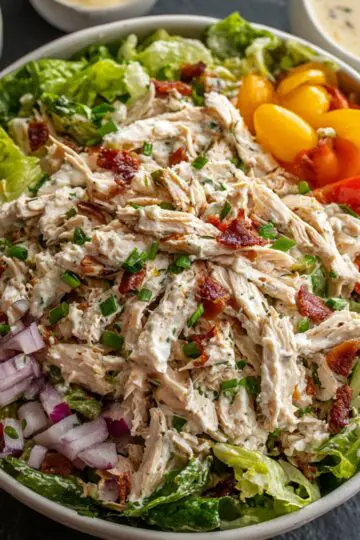 Bowl of creamy Chicken Salad Recipe on a bed of romaine lettuce, topped with shredded chicken, diced red onion, halved cherry tomatoes, and bacon bits, with a small ramekin of dressing and extra bacon in the background.