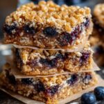 Three homemade oatmeal bars stacked on crumpled parchment over a dark wood surface, with oat flakes and two fresh blueberries beside them, lit by soft side light.