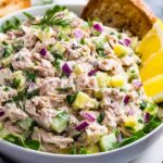 A close-up of a finished tuna salad Recipe in a white bowl, showing flaky tuna mixed with diced celery, chopped red onion, fresh dill, and hard-boiled egg pieces, garnished with lemon wedges and served atop greens.