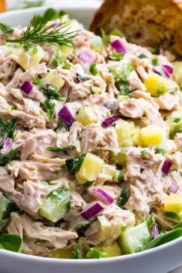 A close-up of a finished tuna salad Recipe in a white bowl, showing flaky tuna mixed with diced celery, chopped red onion, fresh dill, and hard-boiled egg pieces, garnished with lemon wedges and served atop greens.