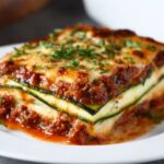 Close-up of a stacked Zucchini Lasagna Recipe slice on a white plate showing alternating layers of zucchini, creamy ricotta, and tomato sauce, topped with melted mozzarella, herbs, and a fresh basil leaf.