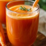 A tall clear glass filled with freshly pressed carrot juice recipe, the bright orange liquid topped with a thin layer of frothy foam. A small sprig of fresh mint or carrot greens floats on the surface, and an unpeeled carrot leans against the glass on a rustic wooden board. In the softly blurred background, another glass of juice and a neutral linen cloth complete the homey scene.