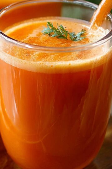 A tall clear glass filled with freshly pressed carrot juice recipe, the bright orange liquid topped with a thin layer of frothy foam. A small sprig of fresh mint or carrot greens floats on the surface, and an unpeeled carrot leans against the glass on a rustic wooden board. In the softly blurred background, another glass of juice and a neutral linen cloth complete the homey scene.