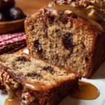 Close-up of a homemade date and walnut cake on a white plate: two thick slices reveal a moist, golden crumb studded with dark date pieces and walnut bits, topped with coarsely chopped nuts and drizzled with caramel-colored sauce, with a softly blurred bowl of dates and a red-and-white tea towel in the background.