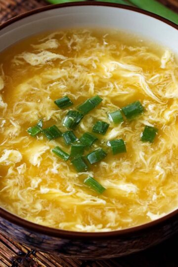 Close-up of a bowl of golden egg drop soup topped with finely chopped green onions, showing delicate silky egg ribbons in clear broth.