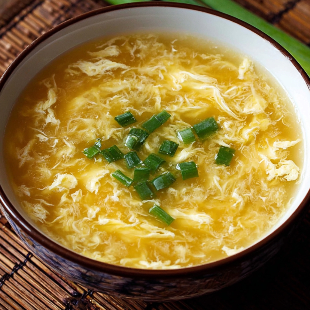 Close-up of a bowl of golden egg drop soup topped with finely chopped green onions, showing delicate silky egg ribbons in clear broth.