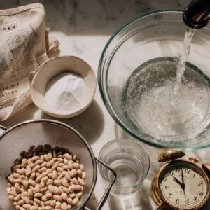 A marble countertop with a glass bowl of clear soaking water, a metal sieve holding pale navy beans, a small dish of baking soda, and a folded linen towel, all arranged around a vintage alarm clock.