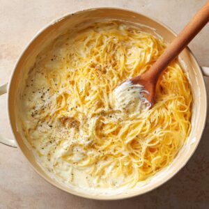 Cooked spaghetti tossed in a creamy white sauce with cracked black pepper, in a large skillet with a wooden spoon.